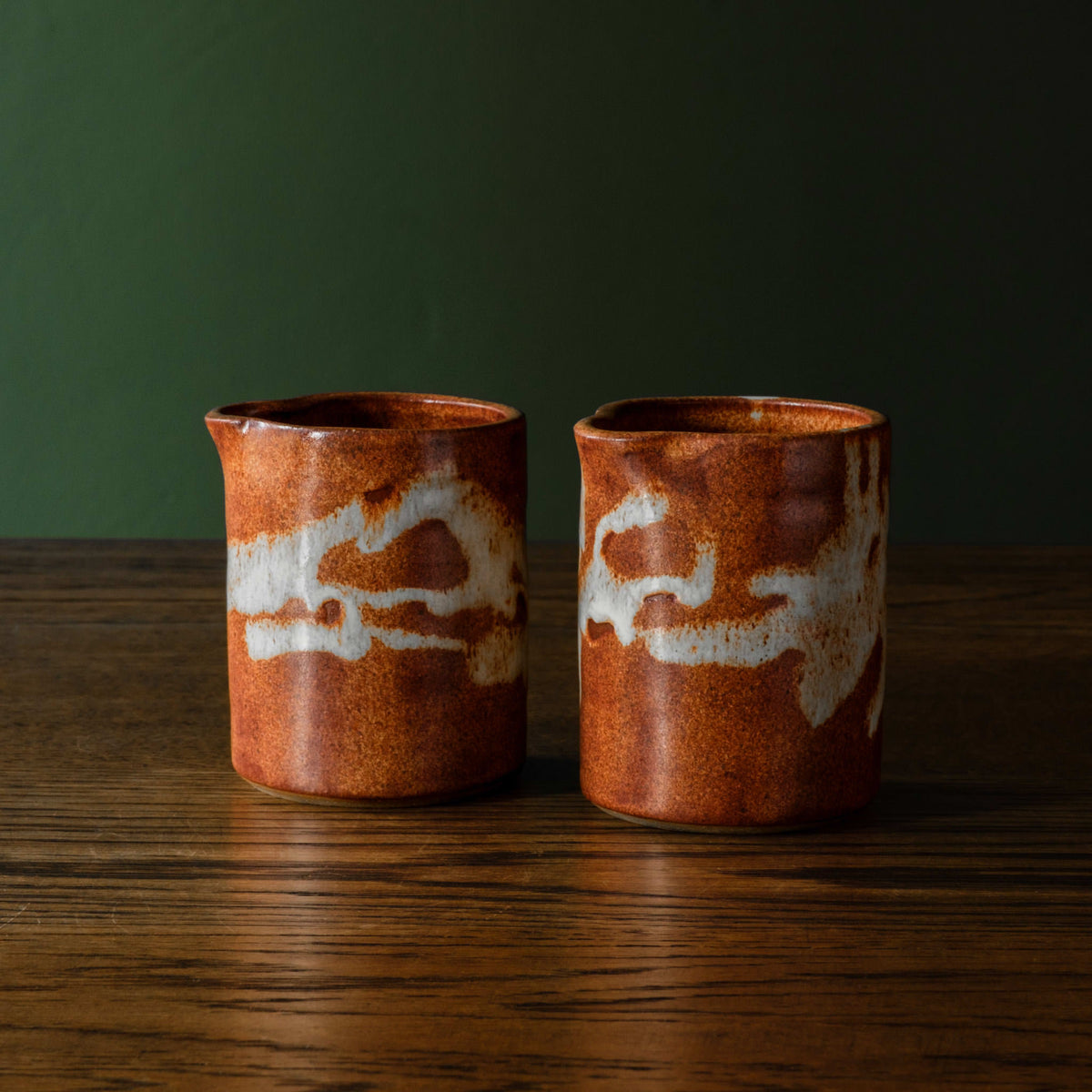 Two small Carrick Ceramics pourers with a glossy terracotta-brown glaze and off-white abstract slip patterns, standing on a dark wooden surface against a dark green background.