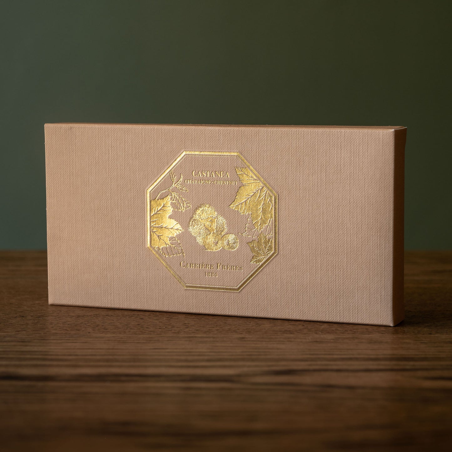 Carrière Frères Chestnut Botanical Palets box. A rectangular textured kraft paper gift box with a gold foil logo on the front, placed on a dark wooden surface against a dark green background.