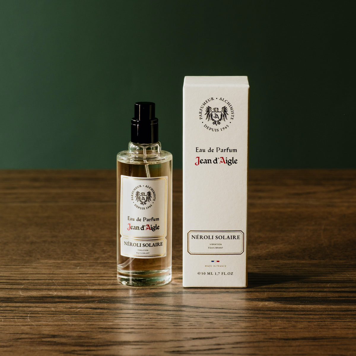 A 50ml bottle of Jean d'Aigle Néroli Solaire Eau de Parfum. In a clear glass bottle with a black spray top beside it’s white product box on a wooden tabletop against a dark green background.