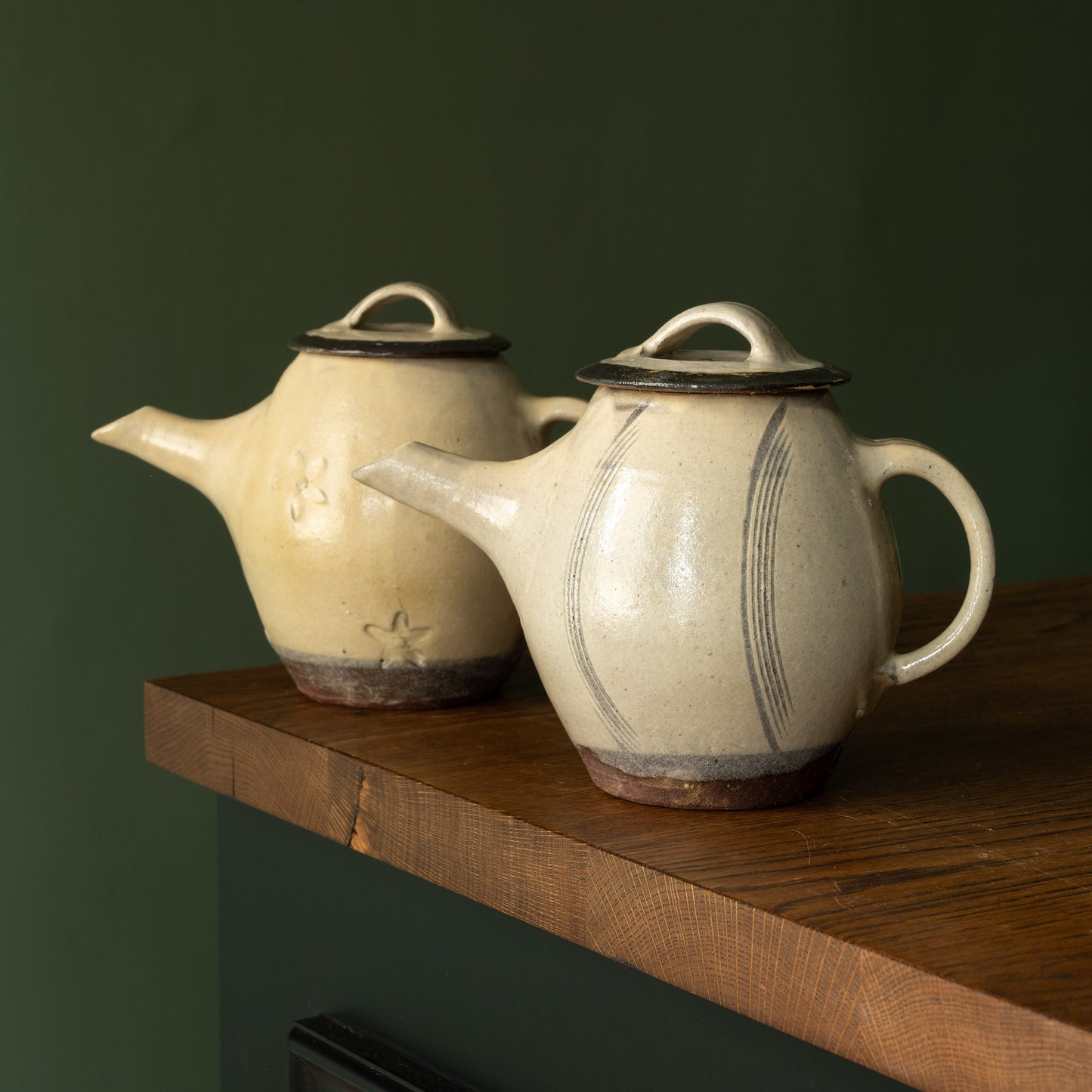 Two Joshua Williams earthenware teapots in a cream glaze on a wooden surface with a dark green background