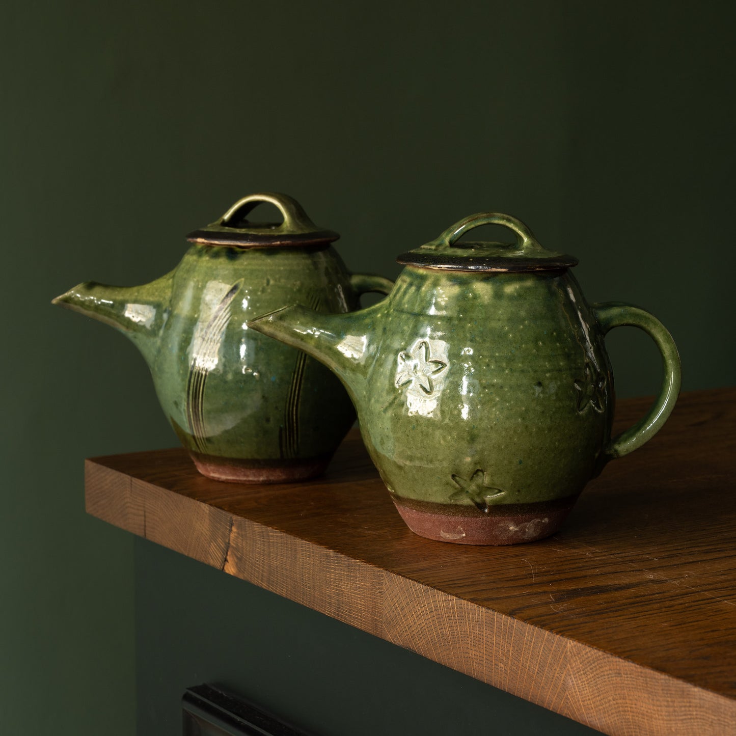 Two Joshua Williams green earthenware teapots on a wooden shelf against a dark green wall.
