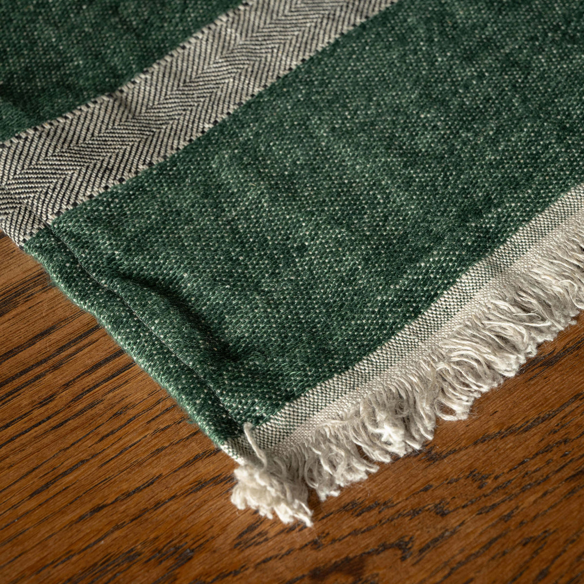 A close-up of Libeco Linen Guest Towel. A dark racing green base with a herringbone patterned border and off-white fringe, laid on a wooden surface.
