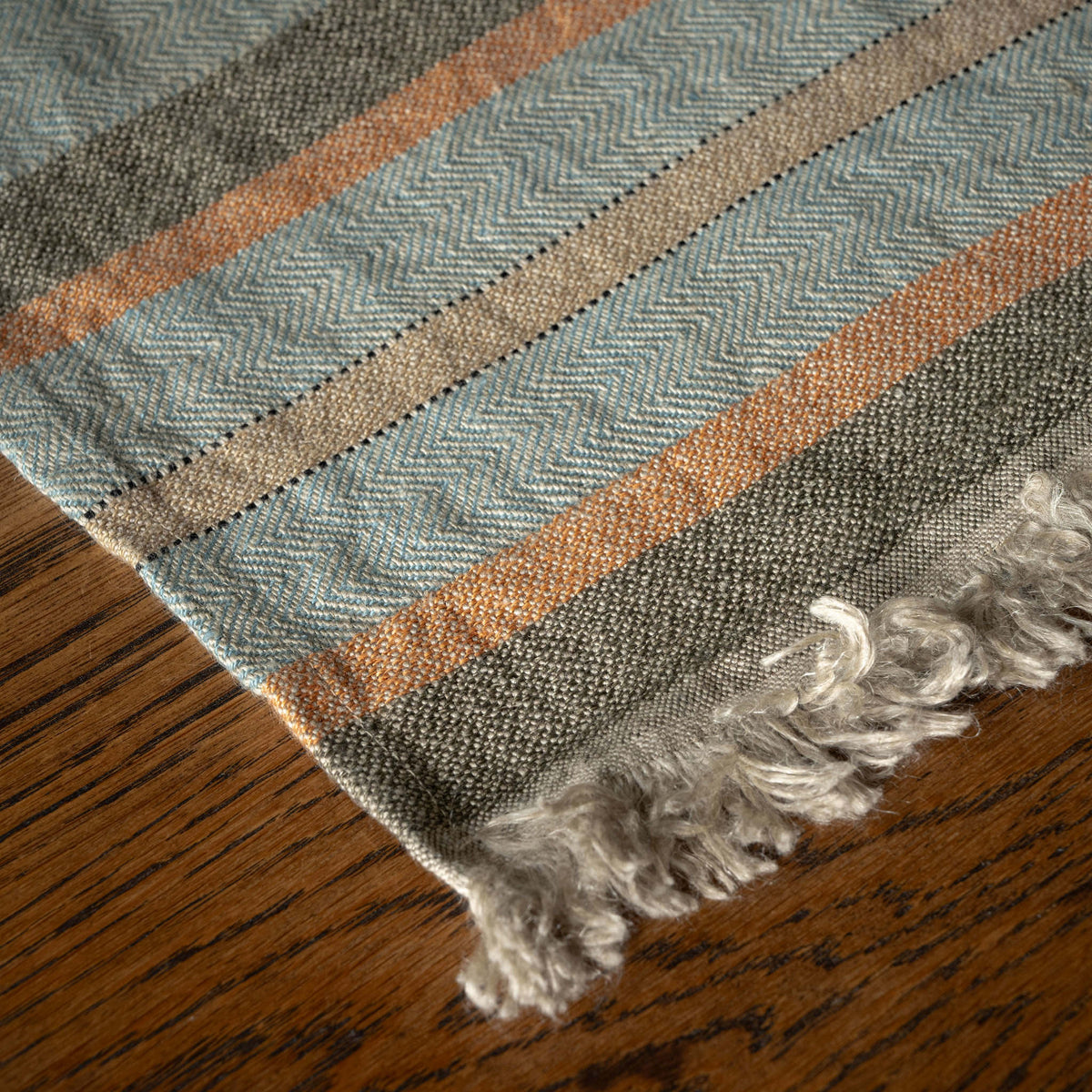 A close-up of Libeco Belgian Linen woven hand towel with a herringbone pattern in muted blue-grey, taupe, and rust orange, finished with fringed edges and shown resting on a wooden surface.
