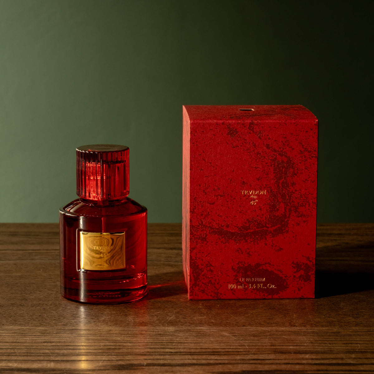 Trudon 45 Eau de Parfum red glass bottle with a gold label stands beside a textured red box on a wooden surface against a dark green background.