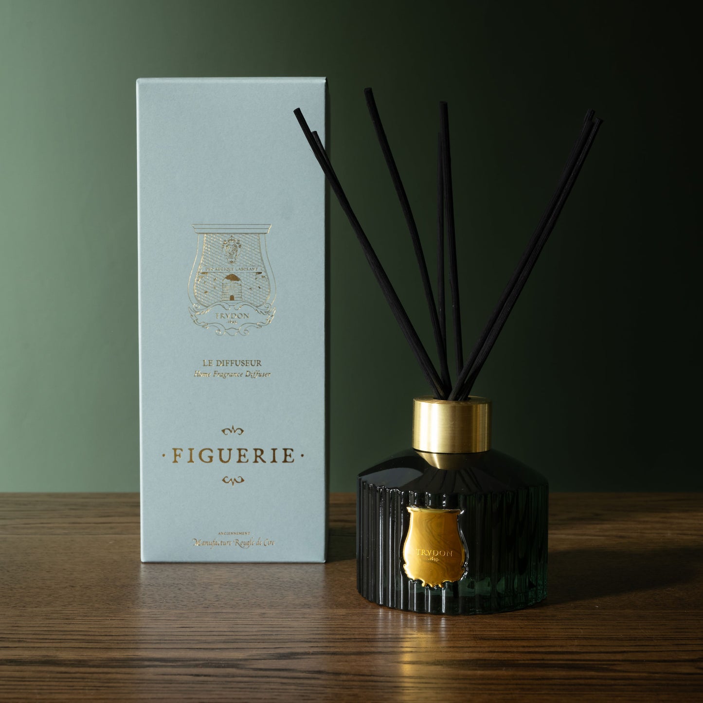 Trudon Figuerie Reed Diffuser with black reeds and gold accents next to its presentation box packaging on a wooden surface.
