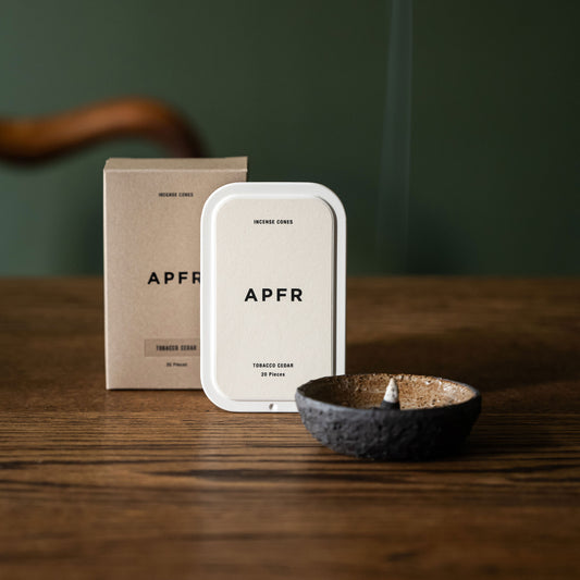 APFR Tobacco Cedar charcoal incense cones & packaging on a wooden table with a small incense bowl with lit incense cone smoking.