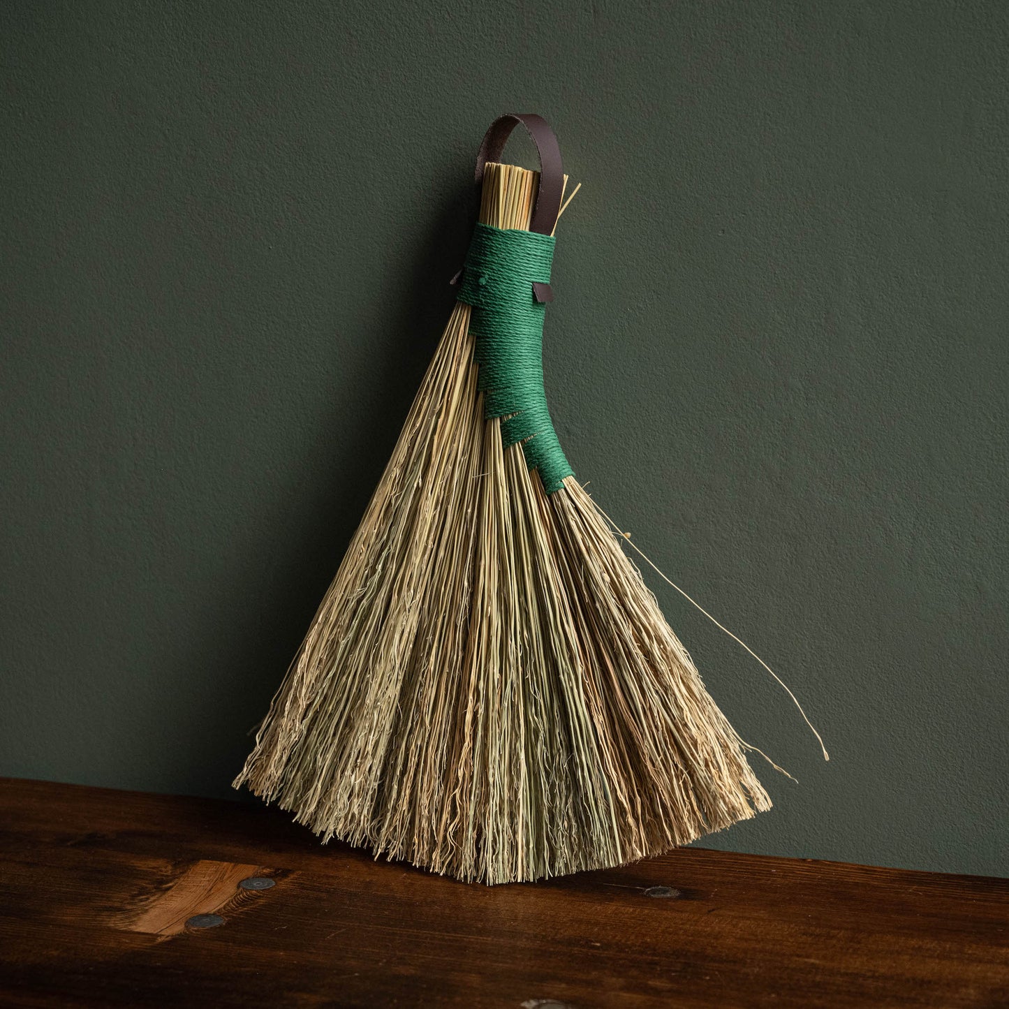 Broomcorn hand broom with green hemp binding and dark brown leather hanging strap. 