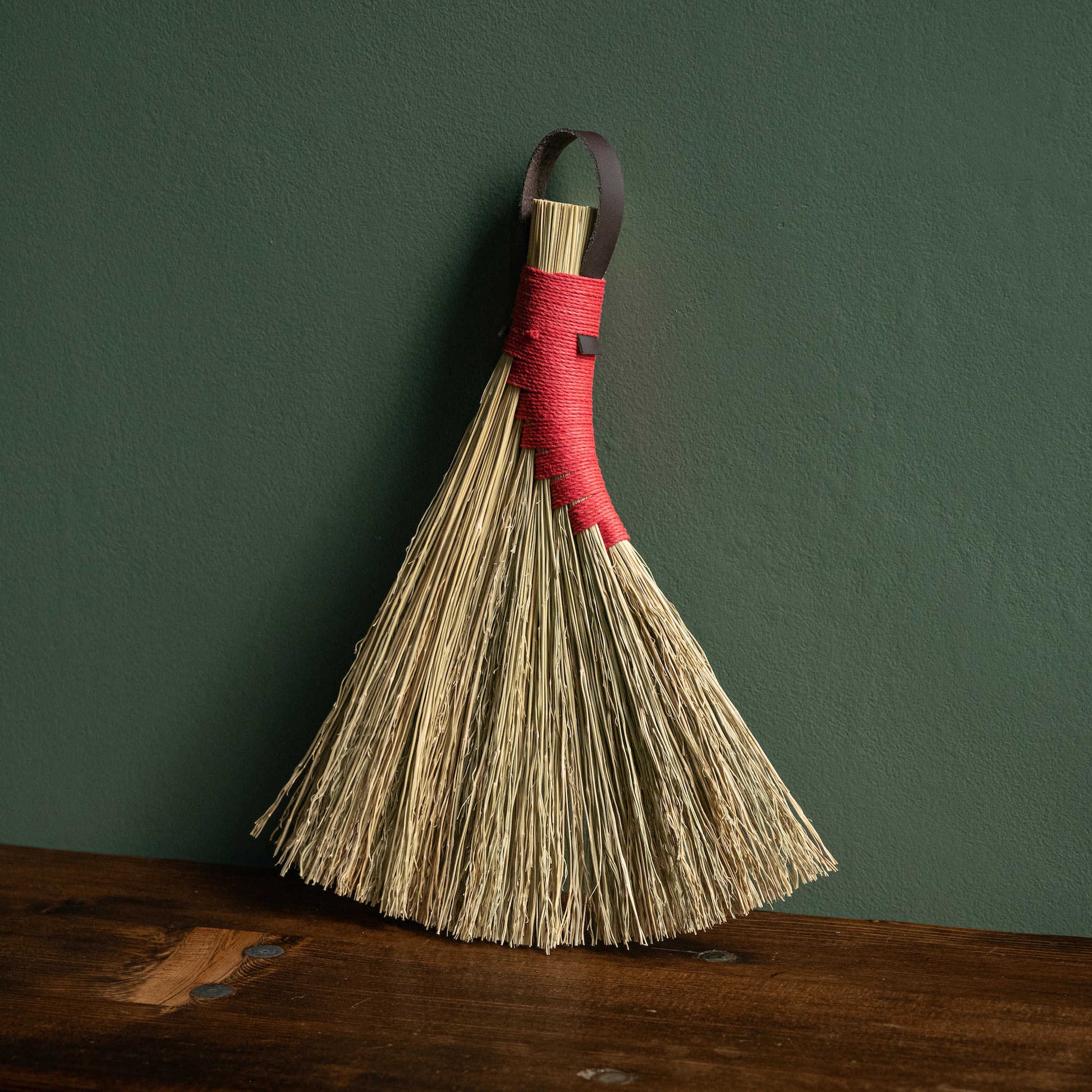 Broomcorn hand broom with red hemp binding and dark brown leather hanging strap. 