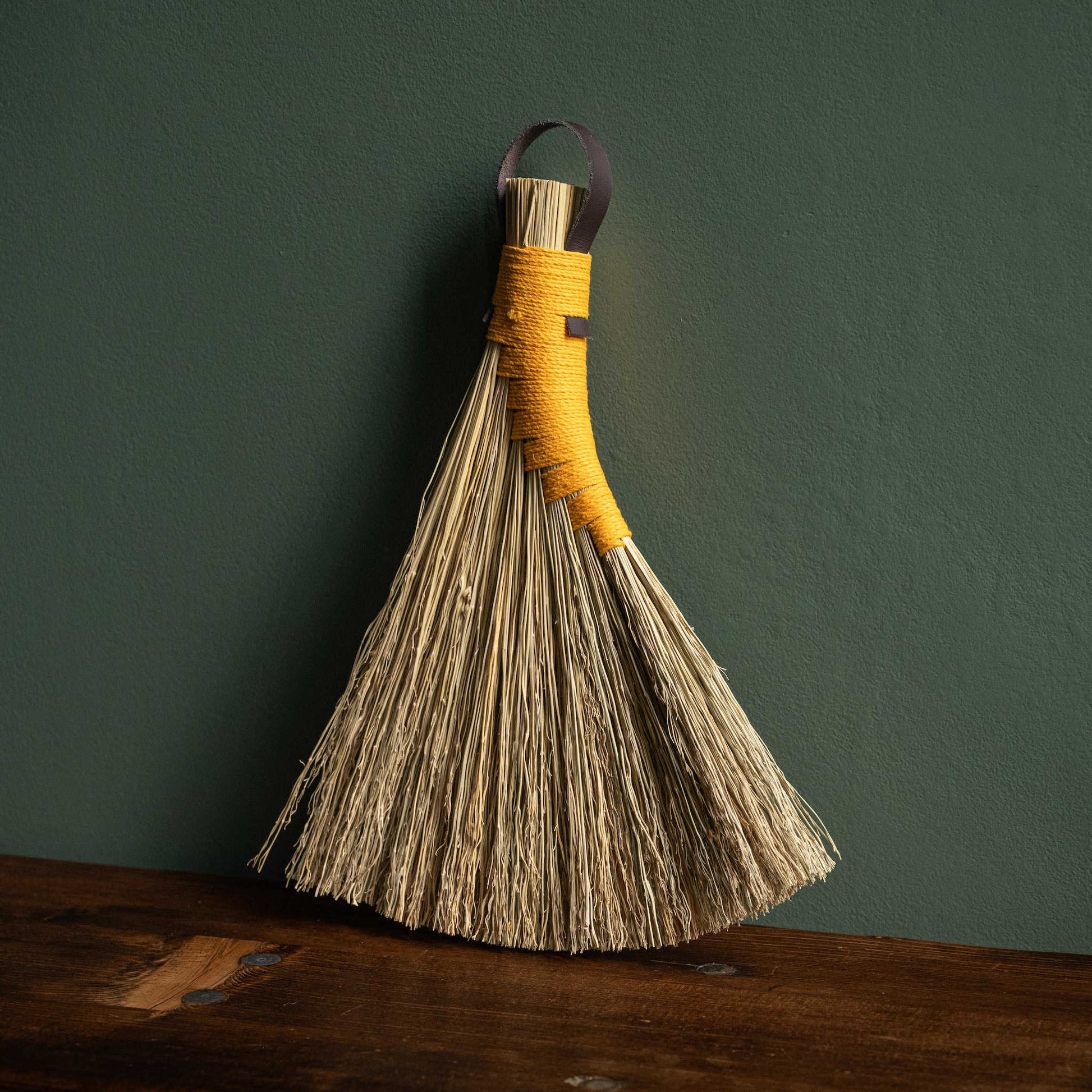 Broomcorn hand broom with yellow hemp binding and dark brown leather hanging strap. 