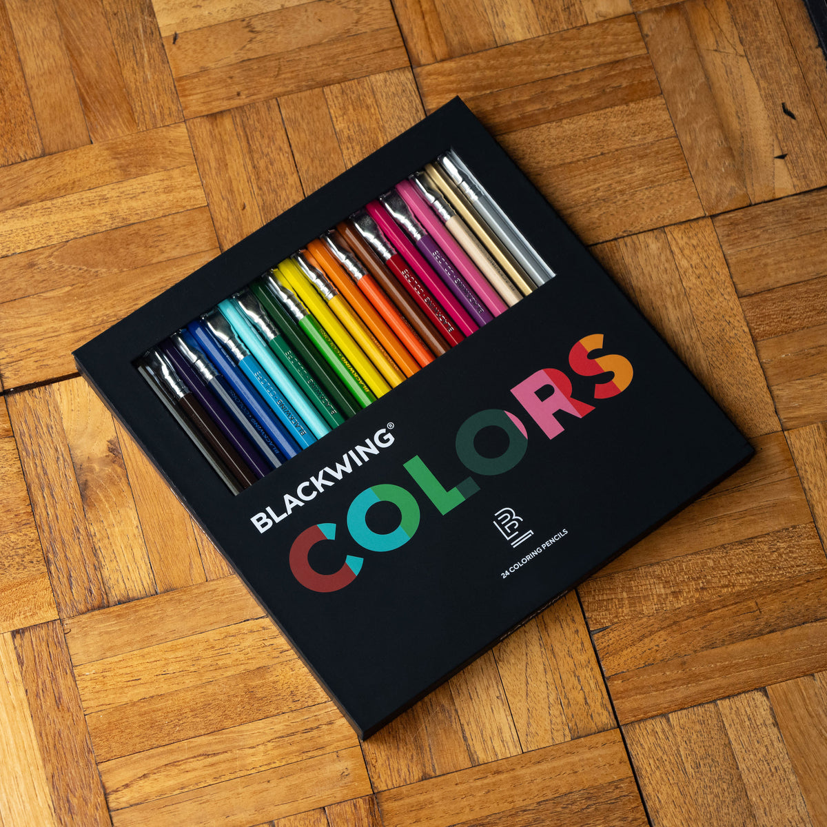 A set of 24 Blackwing Colors drawing pencils in a black box, placed diagonally on a wooden parquet surface.