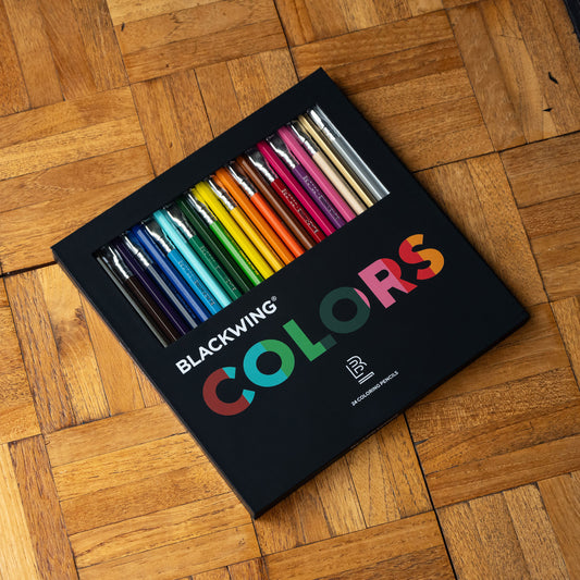 A set of 24 Blackwing Colors drawing pencils in a black box, placed diagonally on a wooden parquet surface.