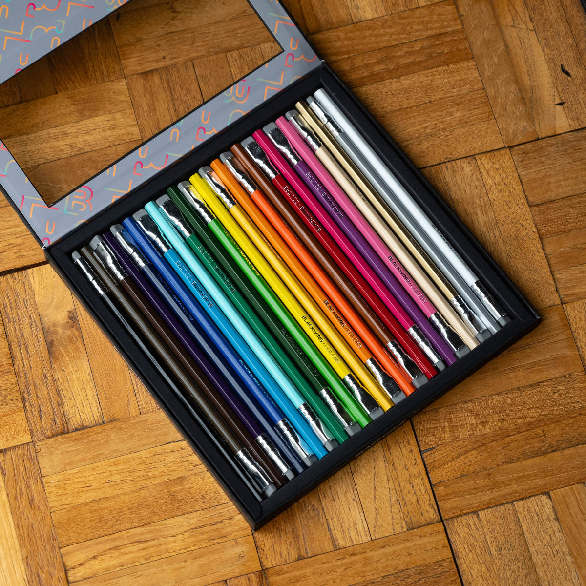 A set of Blackwing colors pencils arranged in a black tray inside a patterned gift box, placed on a wooden parquet surface.