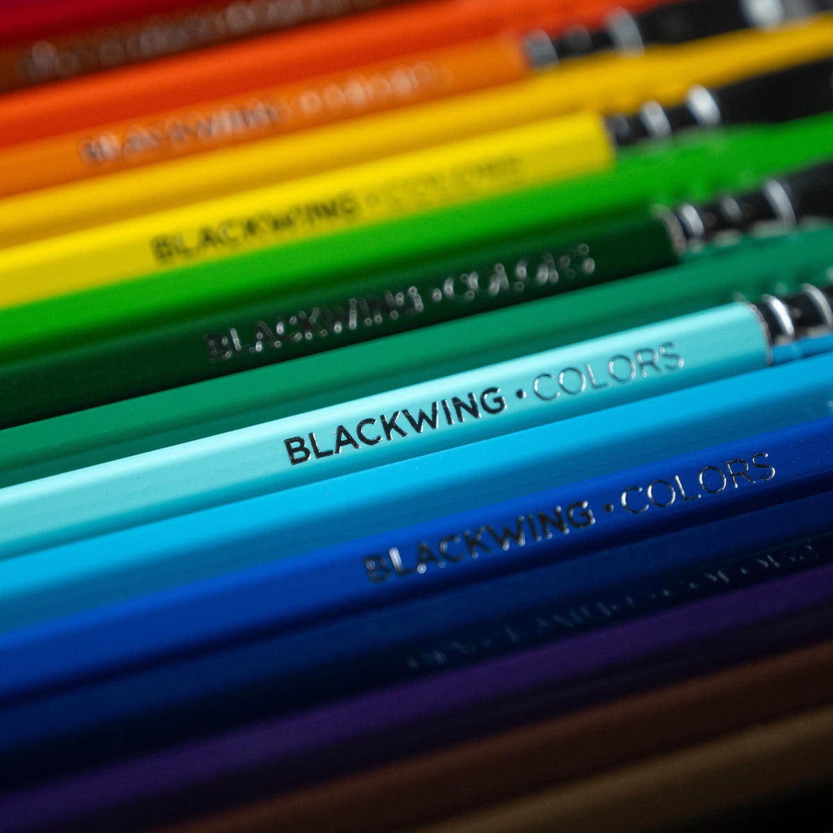 A close-up diagonal view of a set of Blackwing Colors colored pencils in shades of orange, yellow, green, blue, purple, and brown, with the brand name 'BLACKWING COLORS' clearly printed on the barrels.