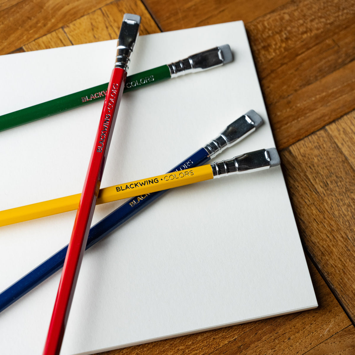 Four Blackwing Colors pencils in red, green, yellow, and blue laid diagonally across a white sketchbook on a wooden table. 