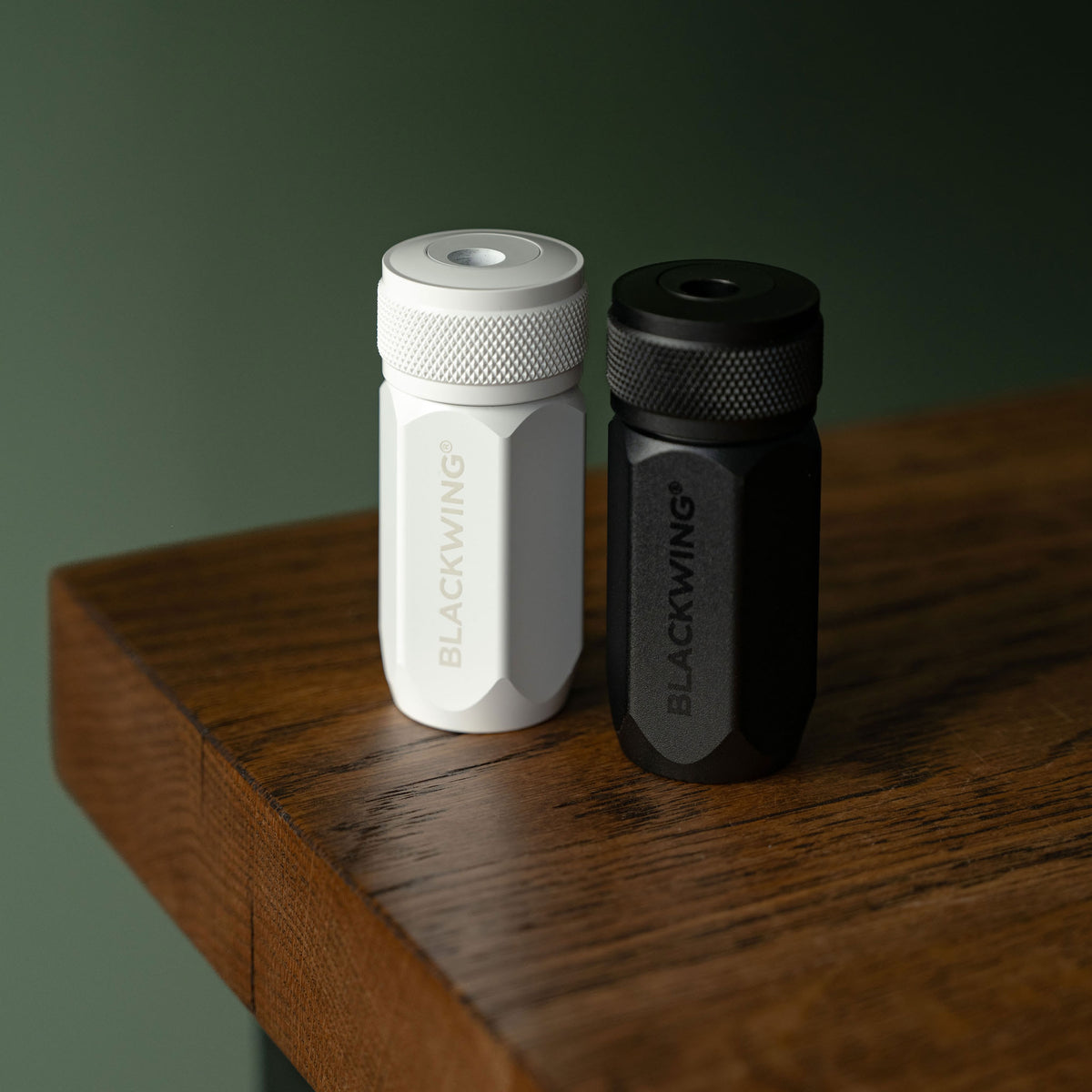 Blackwing Long Point Pencil Sharpeners. Two compact cylindrical accessories with knurled screw-top ends, standing on a wooden tabletop against a dark green wall. One is matte white and the other is matte black, both printed with the visible brand name BLACKWING.