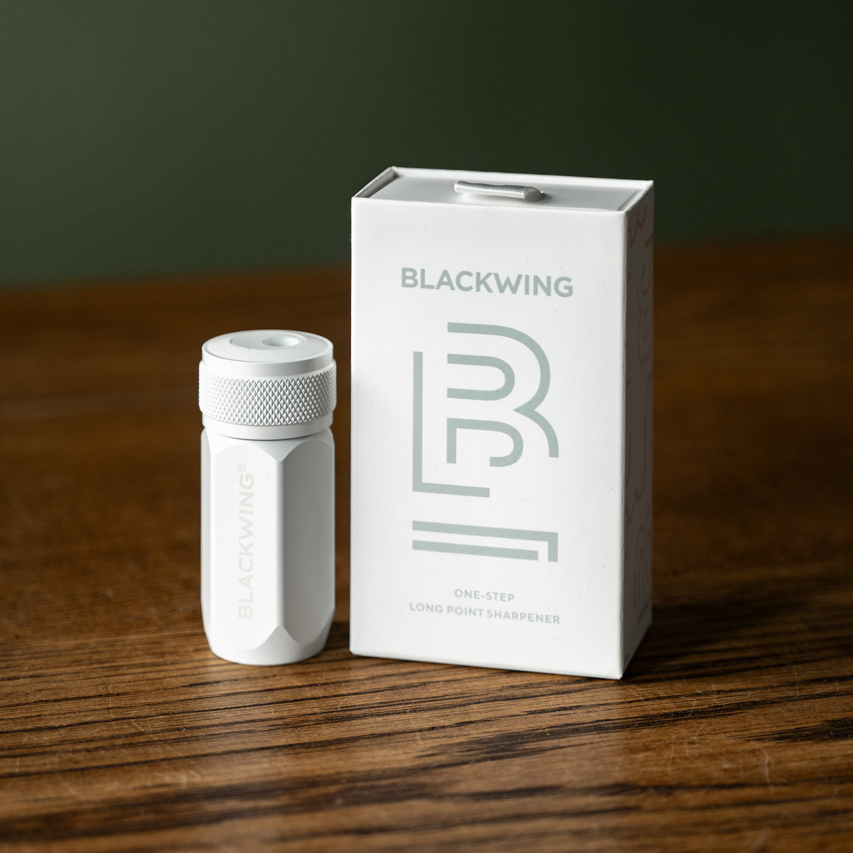 White Blackwing Long Point Pencil Sharpener placed next to its white packaging box on a dark wooden table.