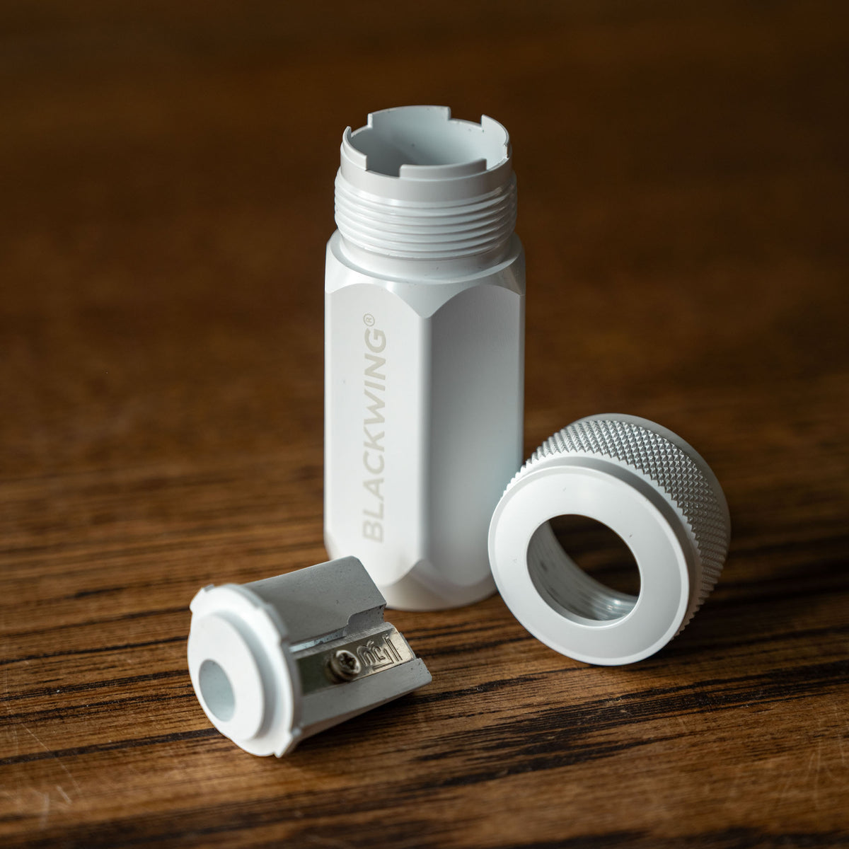 White Blackwing Long Point Pencil Sharpener components showing sharpener blade, screw top and cylindrical body with the brand name BLACKWING printed vertically, shown on a dark wooden surface. 