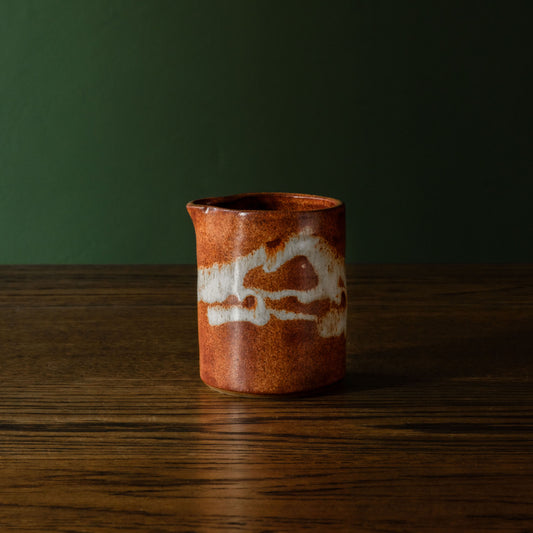 Carrick Ceramics small handmade ceramic pitcher with a rustic brown and rust glaze and an off-white abstract slip design, set on a dark wooden table against a dark green background.