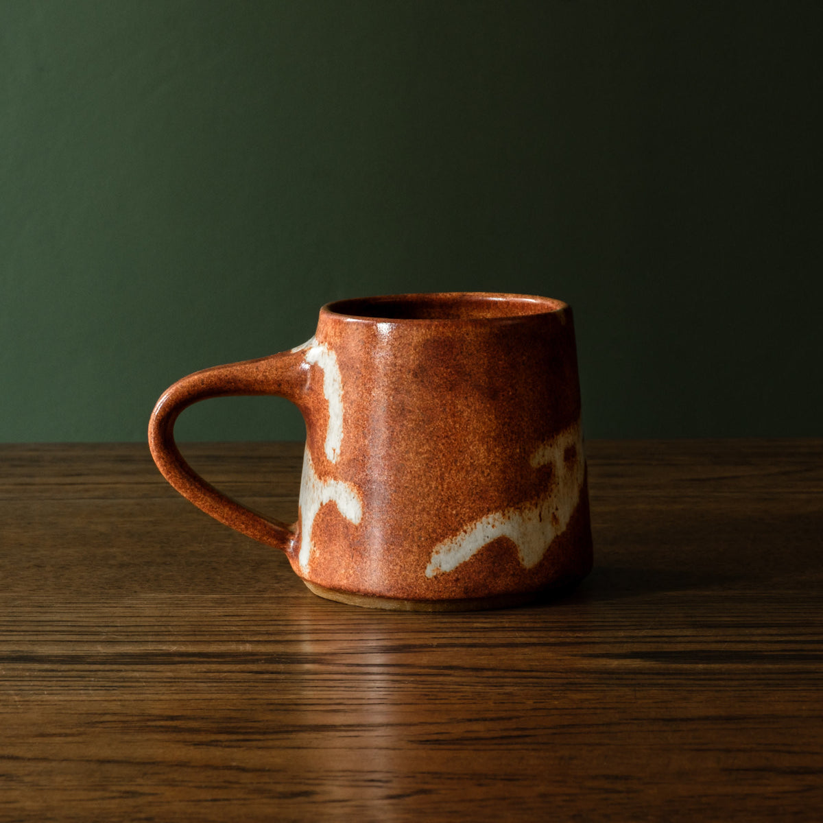 Carrick Ceramics handmade ceramic mug with a warm terracotta brown glaze and off-white abstract accents, sitting on a dark wooden table against a dark green background.