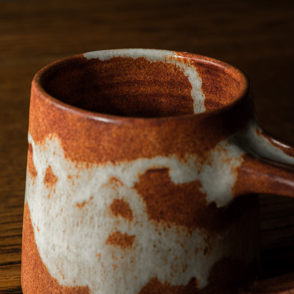 A close-up of Carrick Ceramics  handmade ceramic short mug with a rustic terracotta orange and cream glaze, resting on a dark wooden surface.