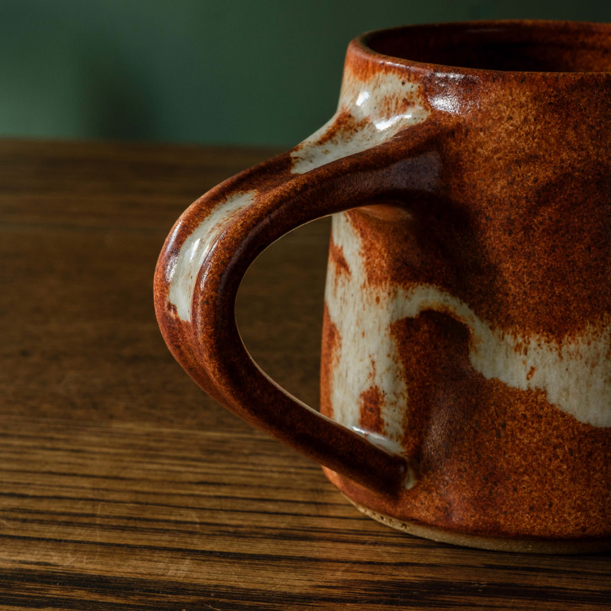 Close up of Carrick Ceramics handmade ceramic mug handle with a warm terracotta brown glaze and off-white abstract accents, sitting on a dark wooden table against a dark green background.