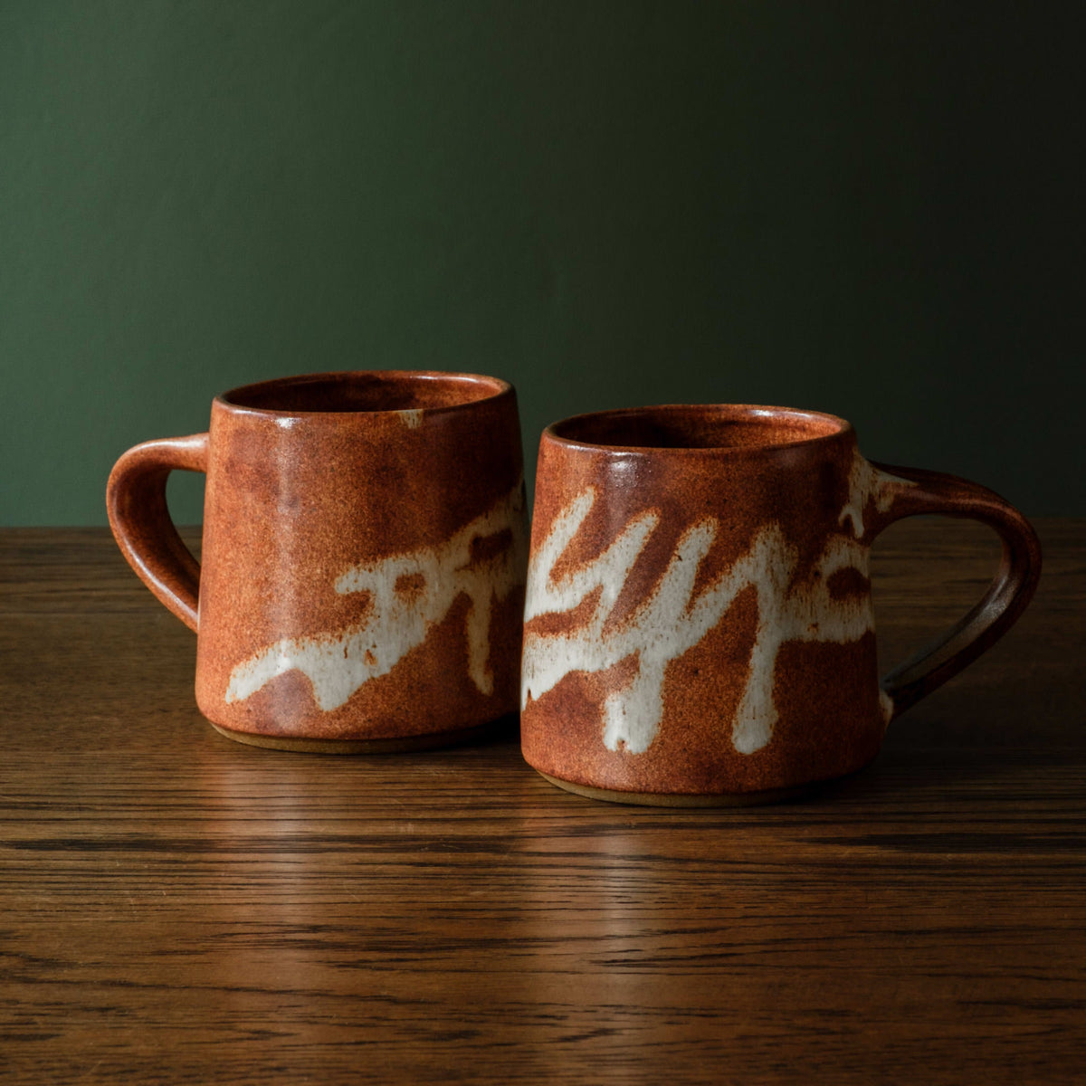 Two Carrick Ceramics handmade ceramic short mugs with a warm terracotta-brown glaze and off-white abstract slip glaze, sitting on a dark wooden table against a dark green background.