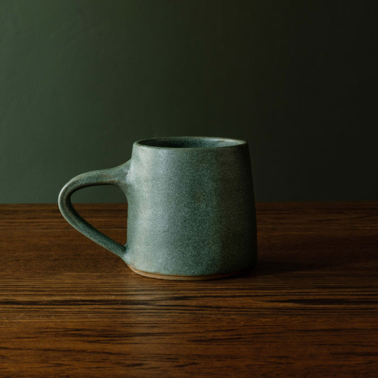Carrick Ceramics handmade ceramic mug with a matt sage green glaze sits on a dark wooden table against a dark olive green background.