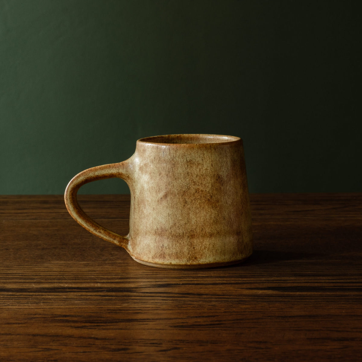 Carrick Ceramics rustic handmade ceramic mug with a textured, earthy brown and beige glaze, sitting on a dark wooden table against a dark green background.