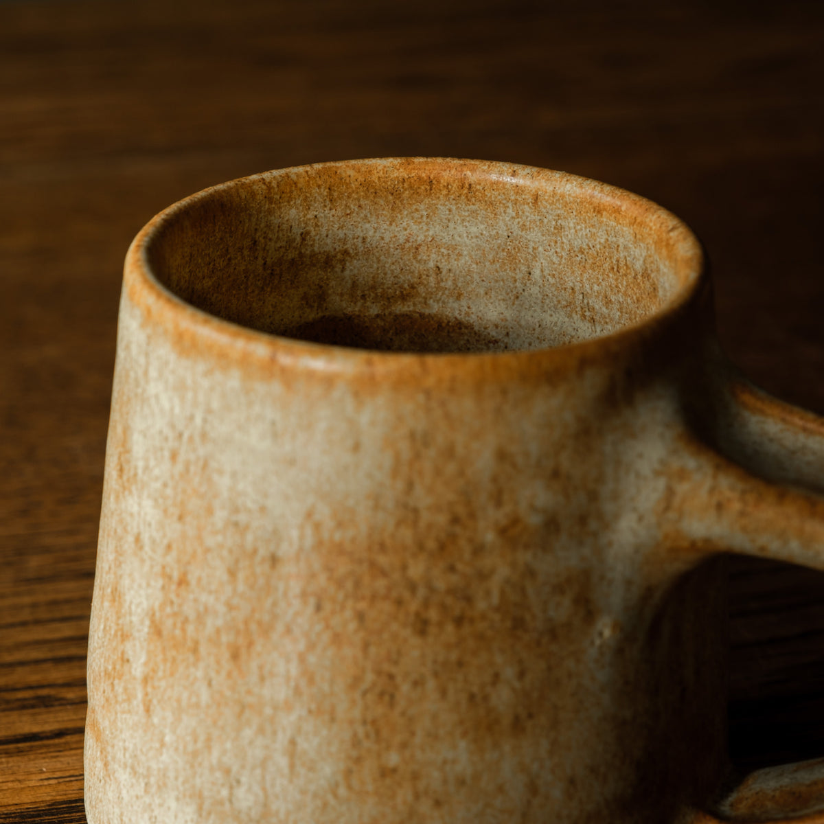 A close-up of Carrick Ceramics rustic handmade ceramic mug with a textured, earthy brown and beige glaze, resting on a dark wooden surface.