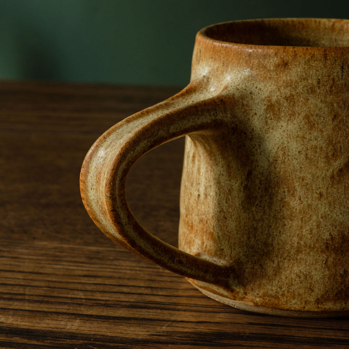A close-up of Carrick Ceramics rustic handmade ceramic mug with a thick, rounded handle and a textured earthy brown and beige glaze, resting on a dark wooden table.