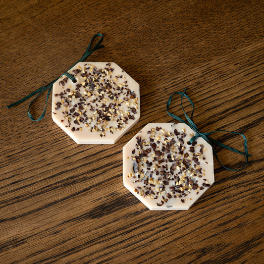 Carrière Frères chestnut scented botanical palets. Two octagonal fragranced wax palets with dried botanicals, tied with dark green ribbons, displayed on a wooden surface.