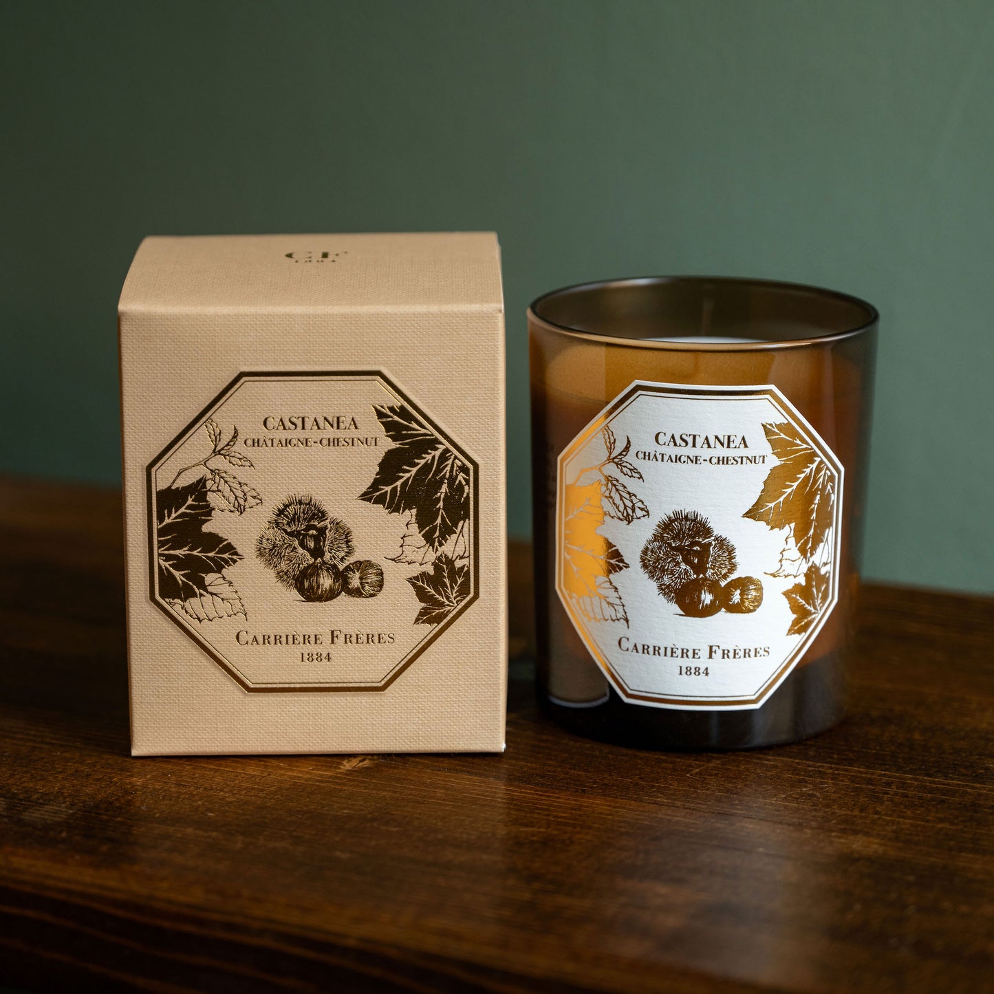 Carrière Frères Chestnut scented candle in an amber glass jar sits on a wooden table beside its cream-coloured product box with botanical illustrations.