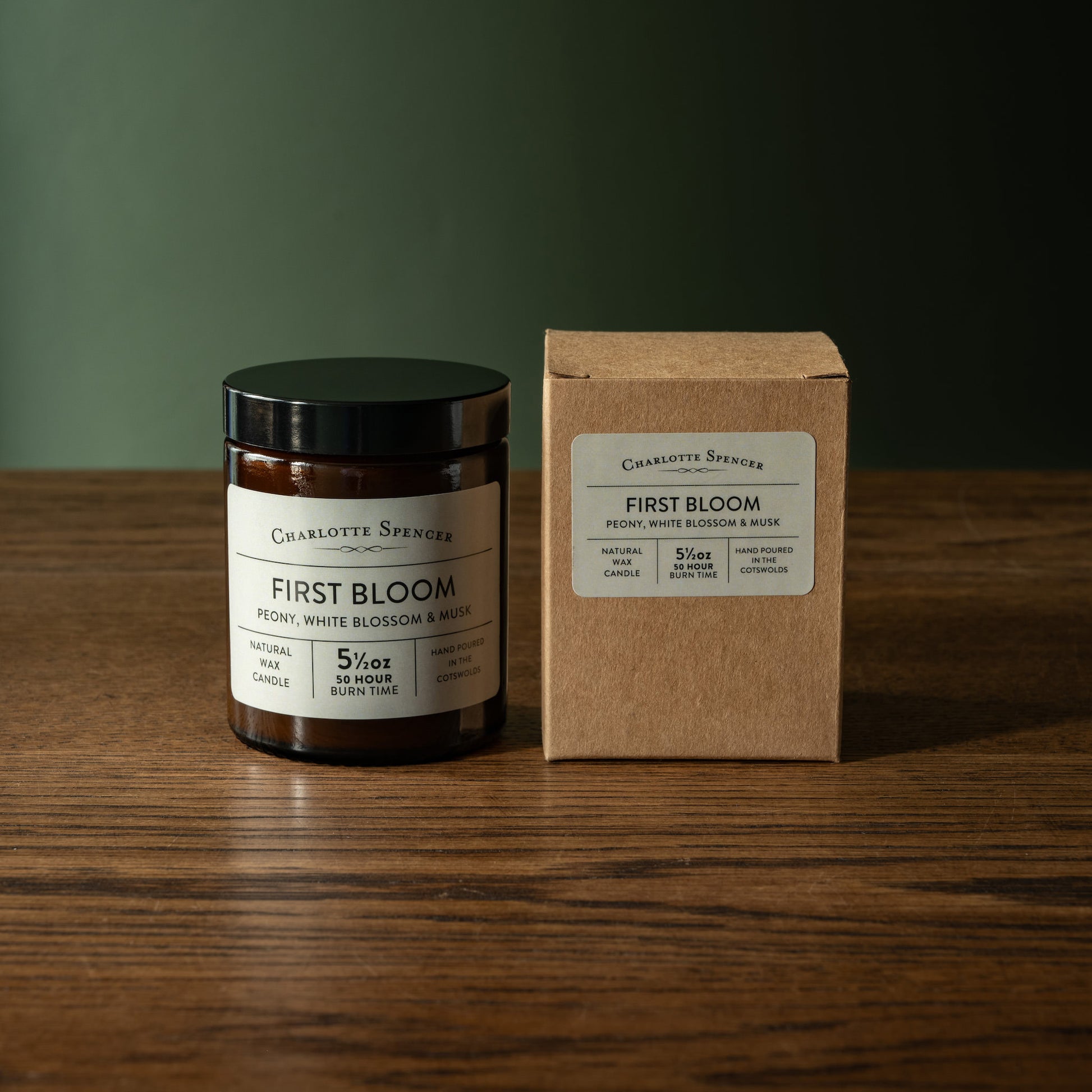 Candle and box of Charlotte Spencer 'First Bloom' candle on a wooden surface with a green background