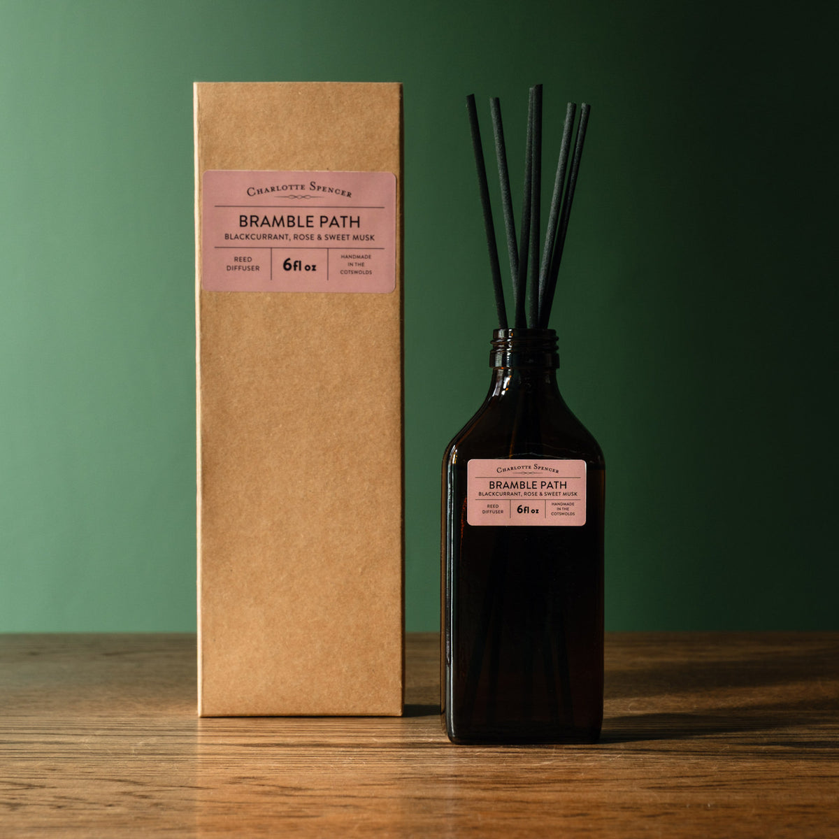 Charlotte Spencer reed diffuser set with a dark amber glass bottle and black reeds, standing beside its brown kraft paper box on a wooden surface against a dark green background. Both pieces feature a branded label reading 'Charlotte Spencer' and 'Bramble Path'.