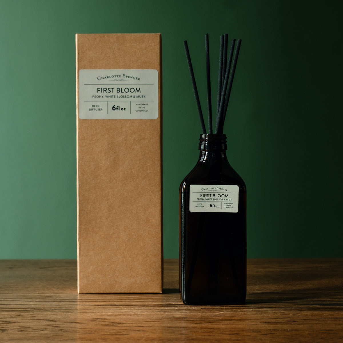 Charlotte Spencer reed diffuser set with a dark amber glass bottle and black reeds, standing beside its brown kraft paper box on a wooden surface against a dark green background. Both pieces feature a branded label reading 'Charlotte Spencer' and 'First Bloom'.