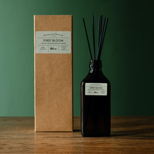 Charlotte Spencer reed diffuser set with a dark amber glass bottle and black reeds, standing beside its brown kraft paper box on a wooden surface against a dark green background. Both pieces feature a branded label reading 'Charlotte Spencer' and 'First Bloom'.