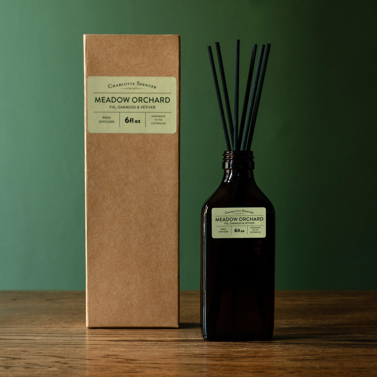 Charlotte Spencer reed diffuser set with a dark amber glass bottle and black reeds, standing beside its brown kraft paper box on a wooden surface against a dark green background. Both pieces feature a branded label reading 'Charlotte Spencer' and 'Meadow Orchard'.