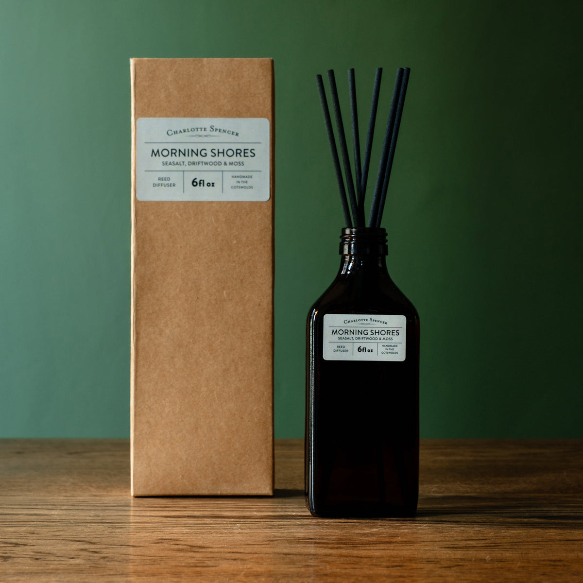 Charlotte Spencer reed diffuser set with a dark amber glass bottle and black reeds, standing beside its brown kraft paper box on a wooden surface against a dark green background. Both pieces feature a branded label reading 'Charlotte Spencer' and 'Morning Shores'.