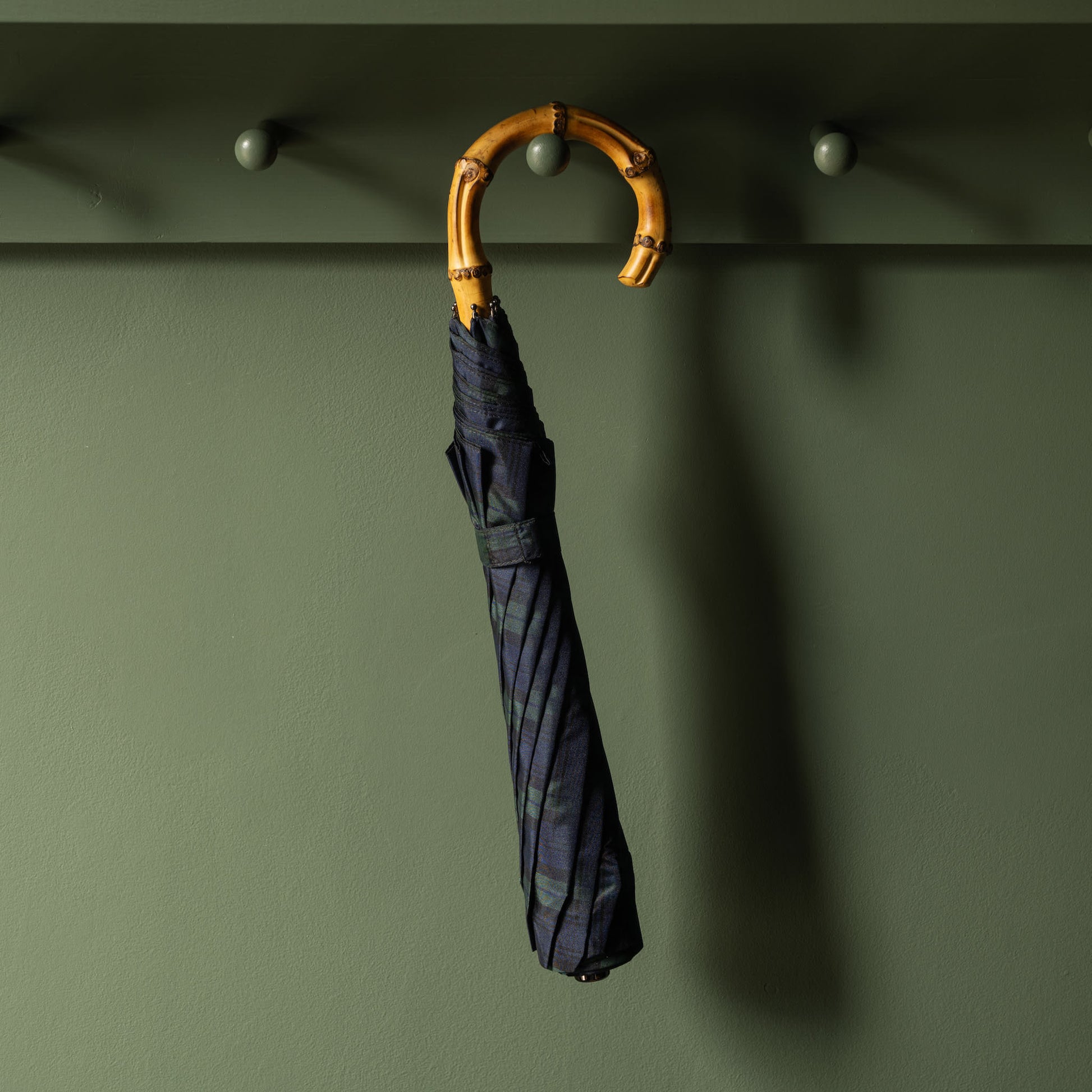 Black Watch Tartan Fox Umbrellas telescopic umbrella with a bamboo handle hanging on a hook against a green wall.