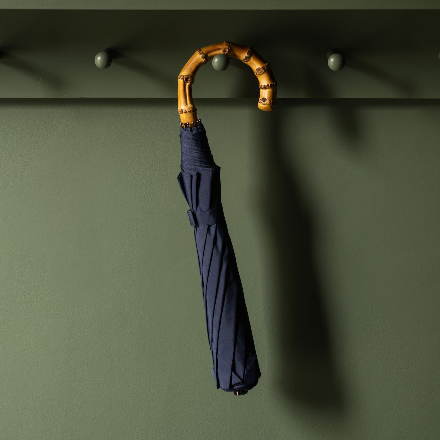 French blue Fox Umbrellas telescopic umbrella with a bamboo handle hanging on a dark green peg rail.