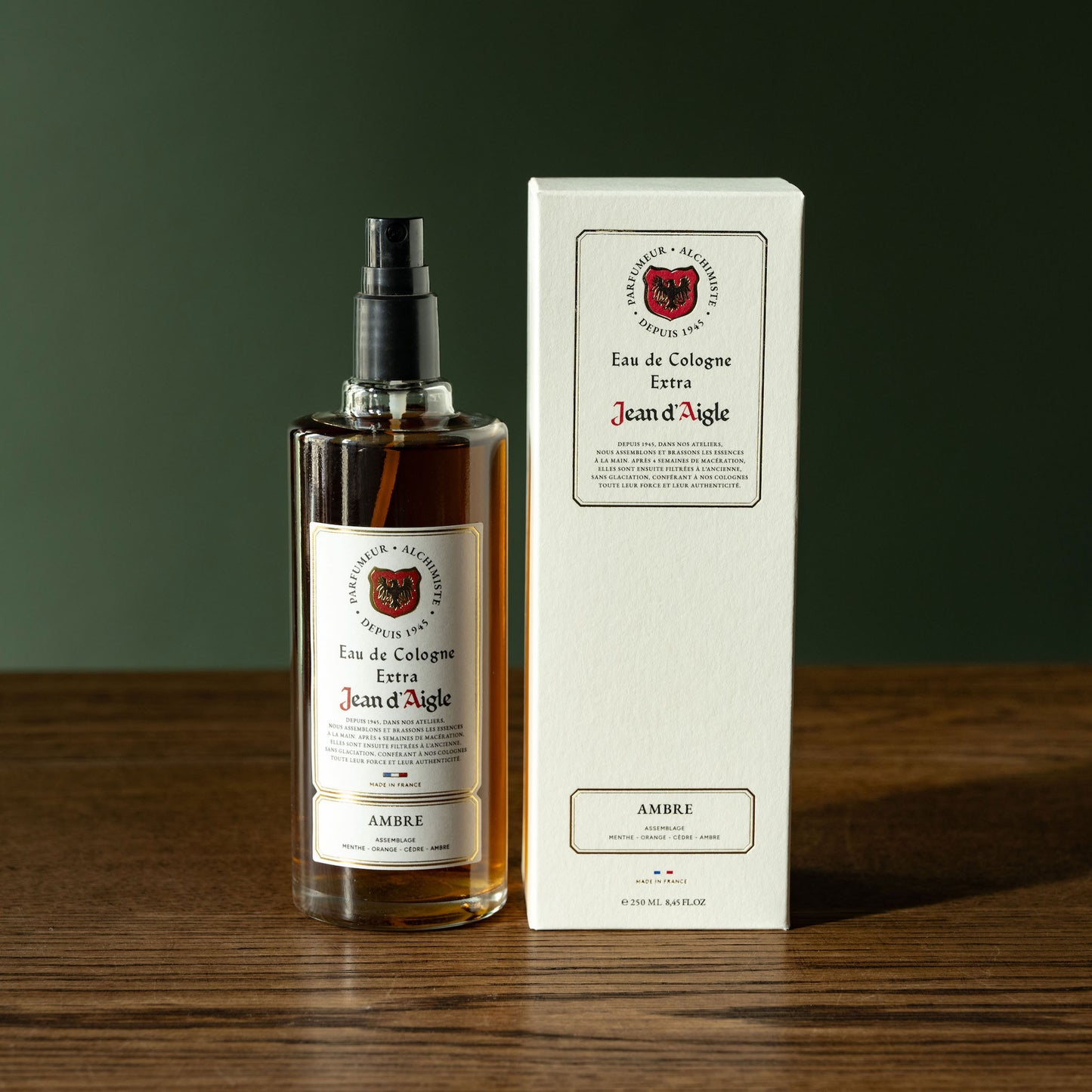 Bottle and box of Jean d'Aigle Ambre Eau de Cologne on a wooden surface with a dark green background