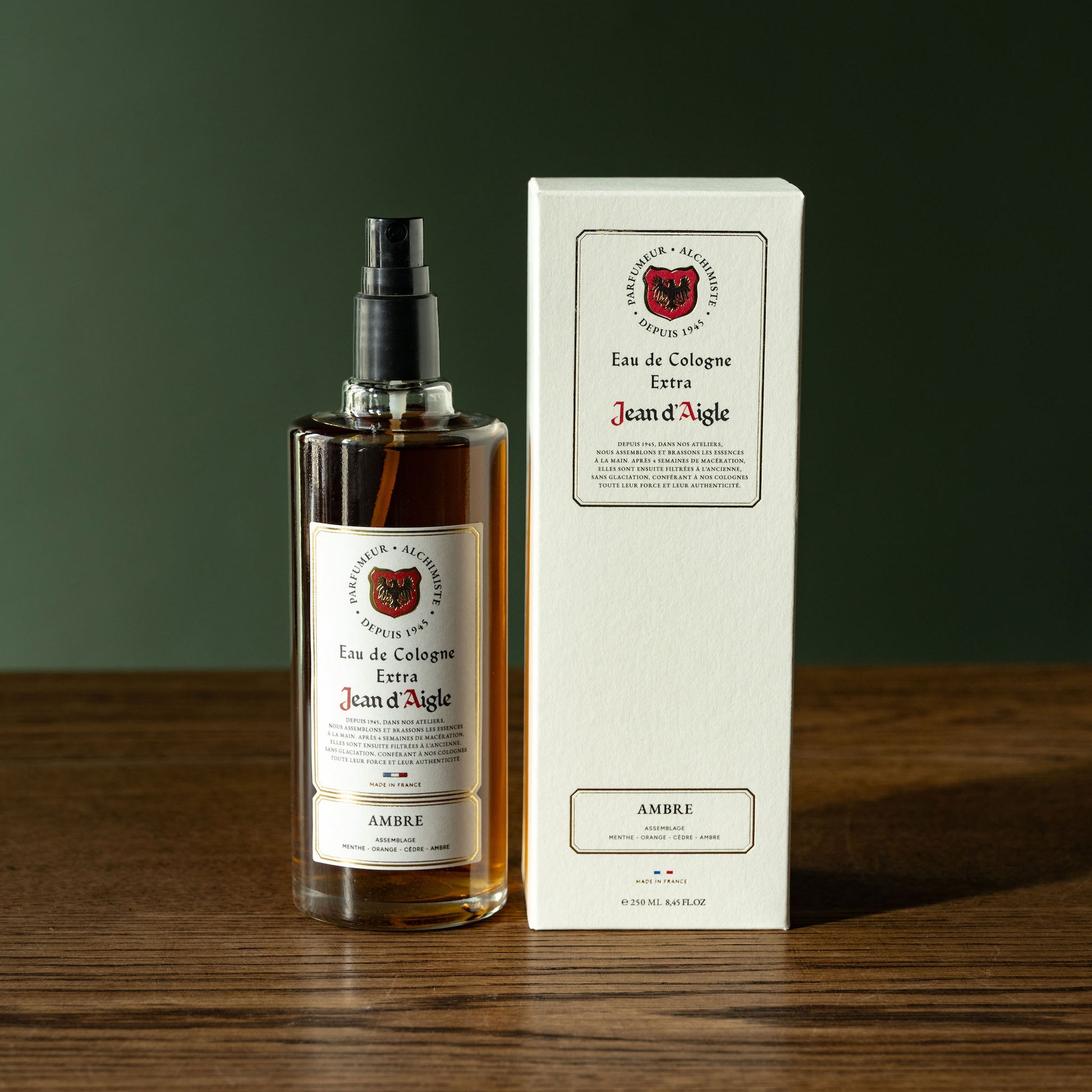 Bottle and box of Jean d'Aigle Ambre Eau de Cologne on a wooden surface with a dark green background