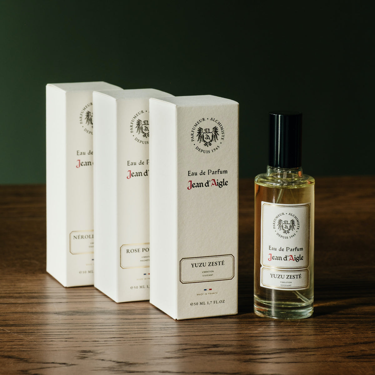 A bottle of Jean d'Aigle Eau de Parfum  next to three boxes of assorted fragrances, arranged on a dark wooden table against a dark green background.