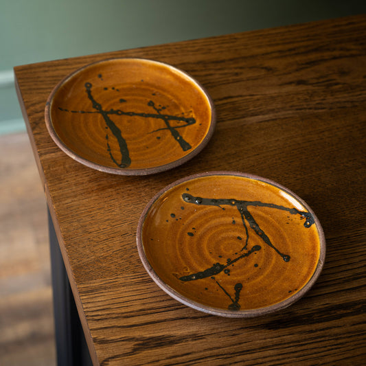Joshua Williams handmade small round earthenware plates with a warm amber-brown glaze and black abstract brushstroke designs, resting on a dark wood tabletop.