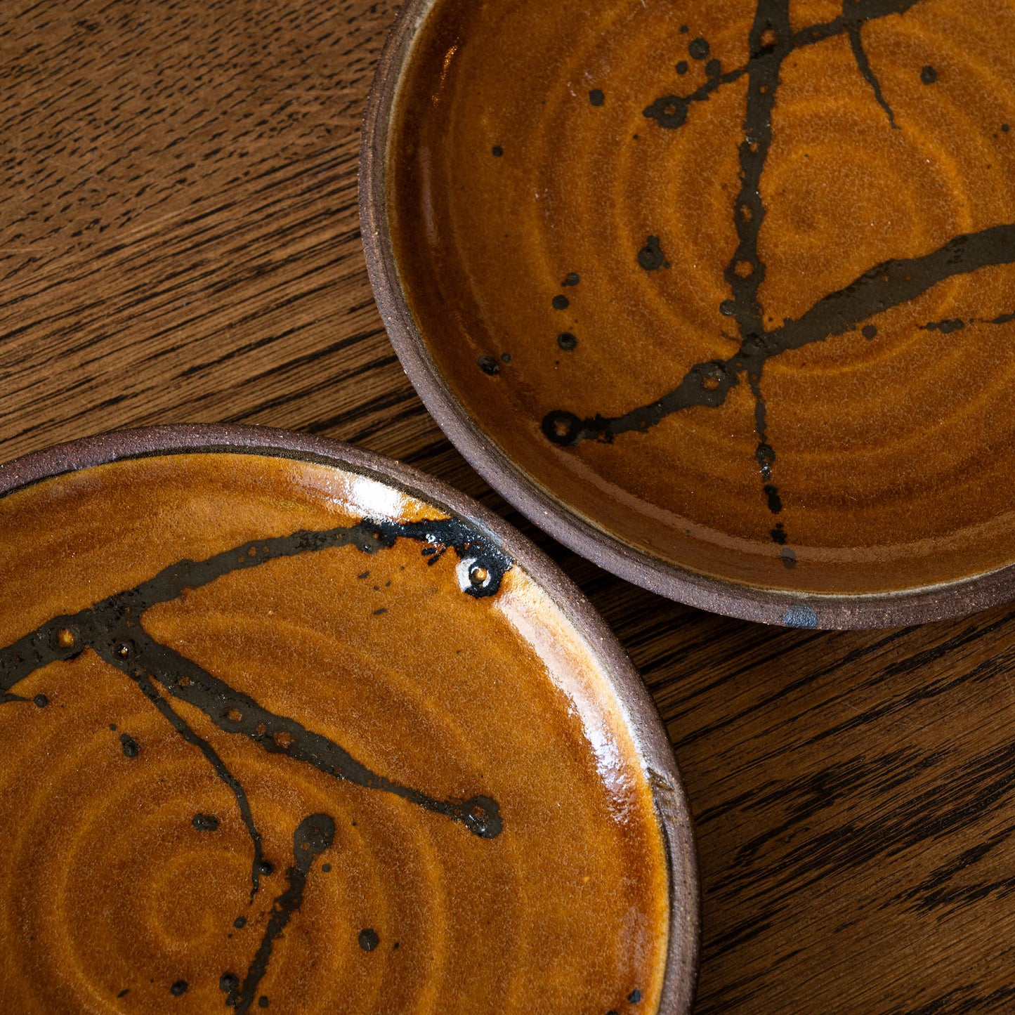 Two handmade Joshua Williams earthenware plates with a glossy amber-brown glaze and dark branch-like slip design brush work, shown on a rustic wooden surface.