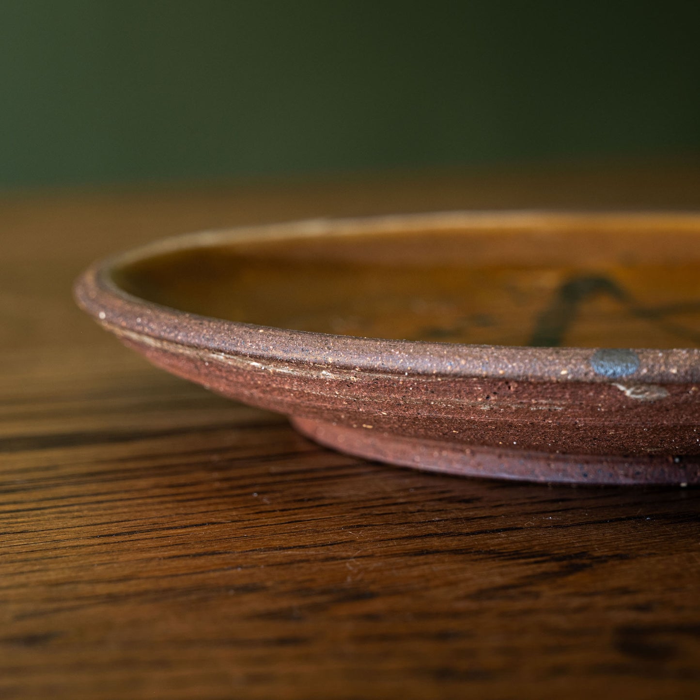 Close-up of a handmade Joshua Williams earthenware shallow plate with a rustic reddish-brown unglazed base, resting on a dark wood tabletop against a dark green background.