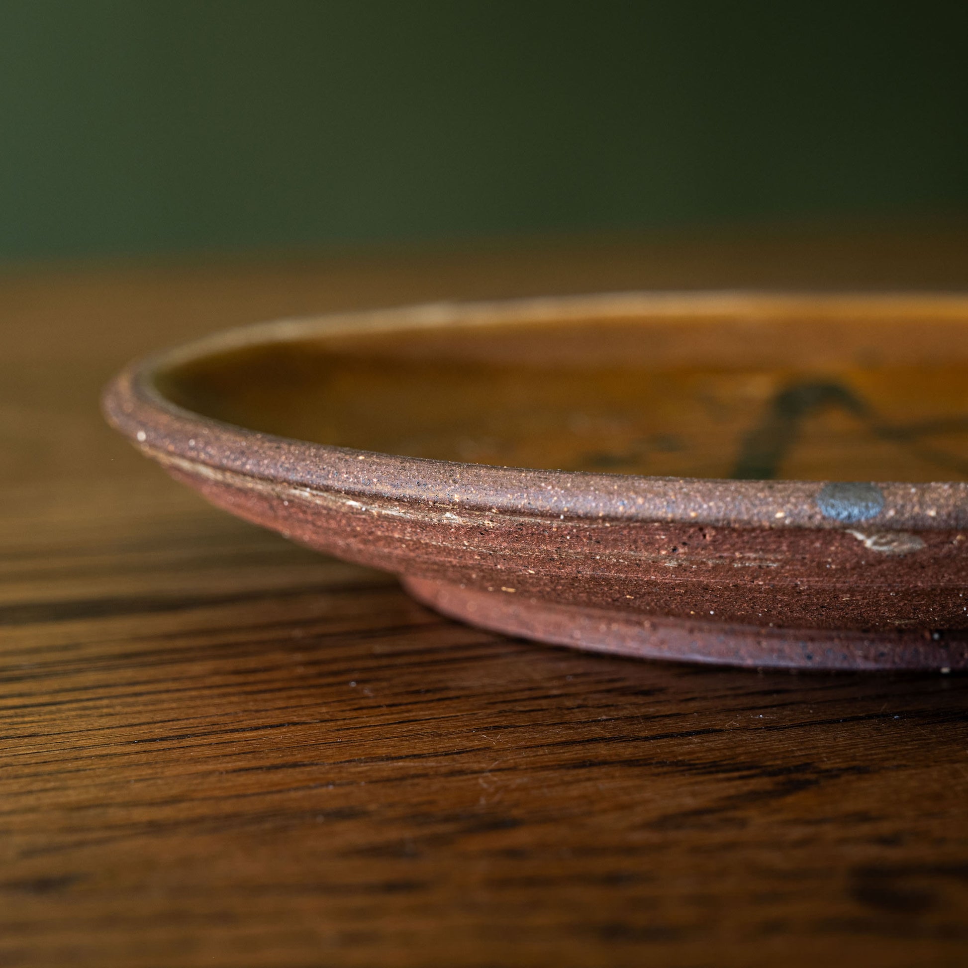 Close-up of a handmade Joshua Williams earthenware shallow plate with a rustic reddish-brown unglazed base, resting on a dark wood tabletop against a dark green background.