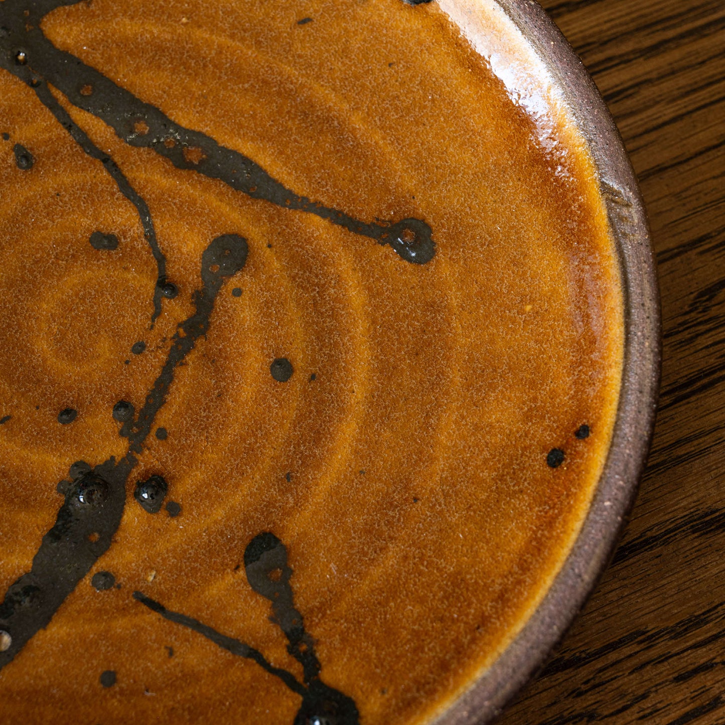 Close-up of Joshua Williams earthenware plate with a warm orange-brown glazed base, with dark brown swirls and splatter design, and a brown rim, set on a dark wood surface.