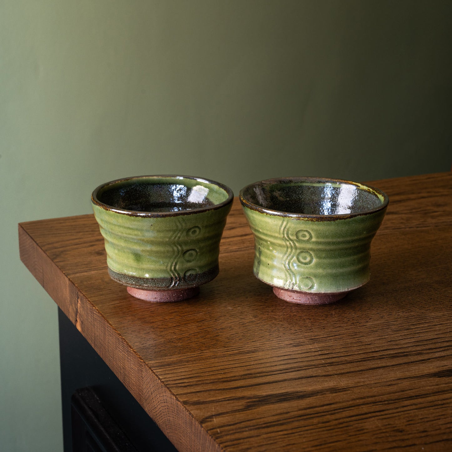 Two handmade Joshua Williams green glazed earthenware tea bowls with a rustic brown base, sitting on a dark wood table against a muted olive green wall.