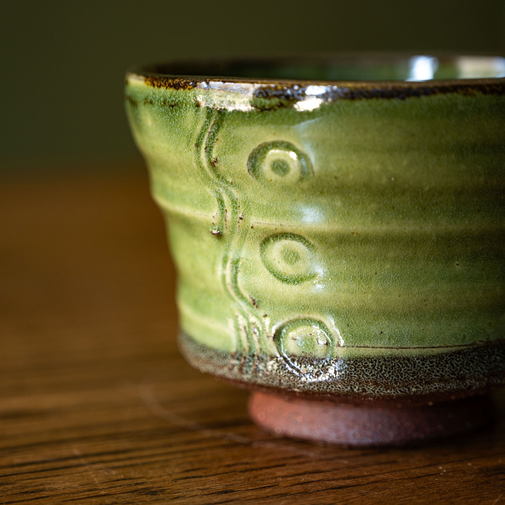 A close-up of a Joshua Williams handmade earthenware tea bowl with a textured dark green glaze, featuring embossed circular patterns and a dark brown unglazed rim, resting on a wooden surface.